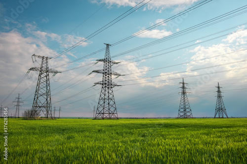 High voltage lines and power pylons in a flat and green agricultural landscape on a sunny day with clouds in the blue sky. Cloudy and rainy. Wheat is growing