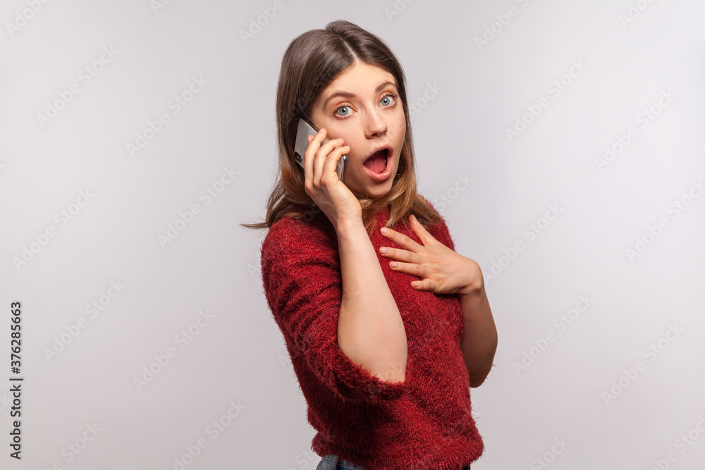 Portrait of astonished brunette woman in shaggy sweater talking with friend on cell phone, looking at camera with surprised expression, shocking rumors. indoor studio shot isolated on gray background Portrait of astonished brunette woman in shaggy sweater talking with friend on cell phone, looking at camera with surprised expression, shocking rumors. indoor studio shot isolated on gray background