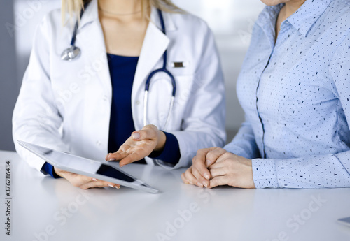Unknown woman-doctor is showing to her patient a description of medication  while sitting together at the desk in the cabinet in a clinic. Female physician is using a computer tablet and a stethoscope