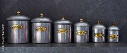 Bild auf Leinwand Set of six pewter jars of different sizes with their lids lined up on a shelf and bearing the inscriptions SUGAR, FLOUR, COFFEE, SPICES, TEA and PEPPER (written in french)