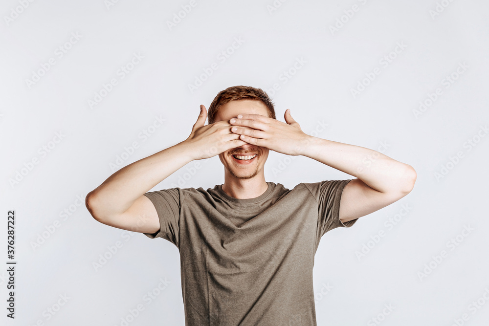 Handsome young man closes his eyes with two hands and smiles. Brunette with a beard in a green shirt posing on a gray background. Place for advertising. I do not see. Handsome young man closes his eyes with two hands and smiles. Brunette with a beard in a green shirt posing on a gray background. Place for advertising. I do not see.