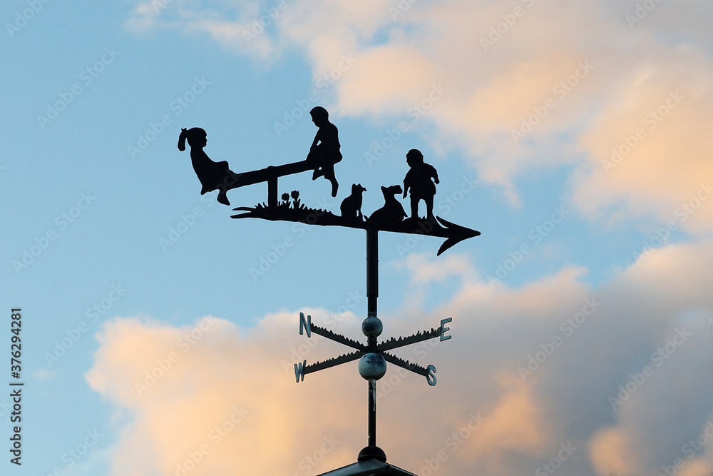Ancient Weathervane with Children Playing Above an Arrow and the Four Cardinal directions of East, West, North and South on a Blue Sky Background Ancient Weathervane with Children Playing Above an Arrow and the Four Cardinal directions of East, West, North and South on a Blue Sky Background