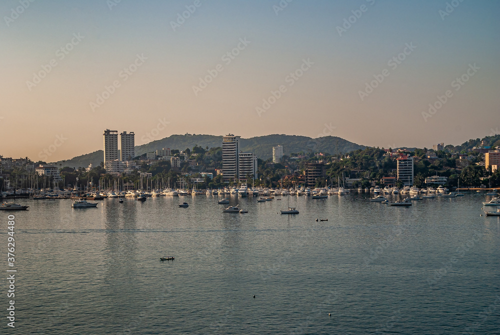 Acapulco, Mexico - November 25, 2008: Twilihgt over the blue water bay with yacht harbor and highrise buildings along shoreline with highrise buildings. Backdrop of green hills, under light blue sky.