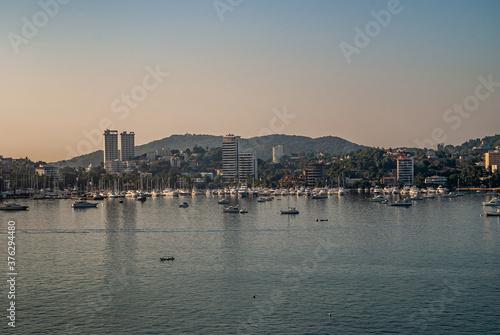 Acapulco, Mexico - November 25, 2008: Twilihgt over the blue water bay with yacht harbor and highrise buildings along shoreline with highrise buildings. Backdrop of green hills, under light blue sky.