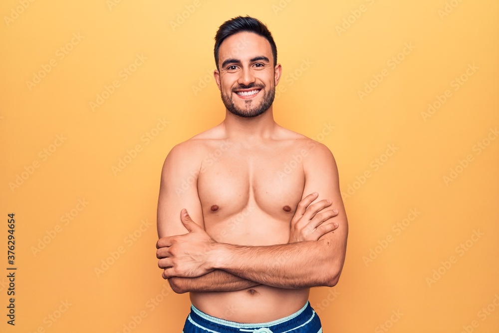Young handsome man with beard wearing sleeveless t-shirt standing over yellow background happy face smiling with crossed arms looking at the camera. Positive person.