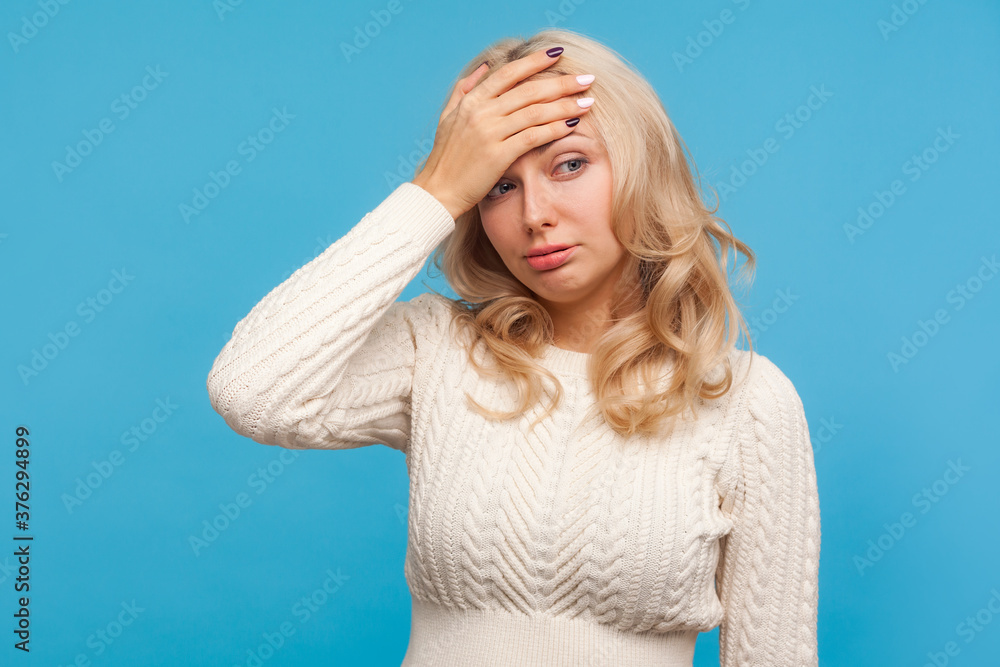 Depressed hopeless woman with curly blond hair making facepalm gesture, shame, getting into trouble, making mistake. Indoor studio shot isolated on blue background
