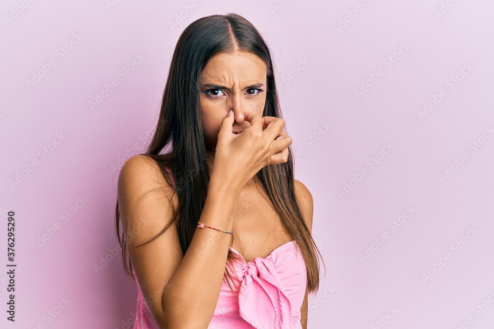 Young hispanic woman wearing casual style with sleeveless shirt smelling something stinky and disgusting, intolerable smell, holding breath with fingers on nose. bad smell Young hispanic woman wearing casual style with sleeveless shirt smelling something stinky and disgusting, intolerable smell, holding breath with fingers on nose. bad smell
