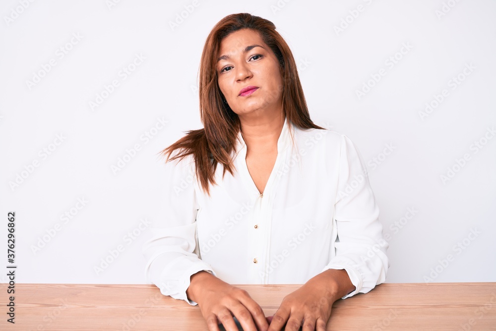 Middle age brunette hispanic business woman wearing casual white shirt sitting on the table relaxed with serious expression on face. simple and natural looking at the camera. Middle age brunette hispanic business woman wearing casual white shirt sitting on the table relaxed with serious expression on face. simple and natural looking at the camera.