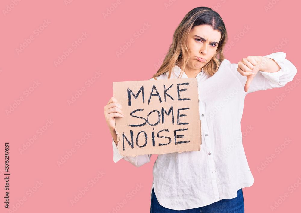 Young caucasian woman holding make some noise banner with angry face, negative sign showing dislike with thumbs down, rejection concept Young caucasian woman holding make some noise banner with angry face, negative sign showing dislike with thumbs down, rejection concept