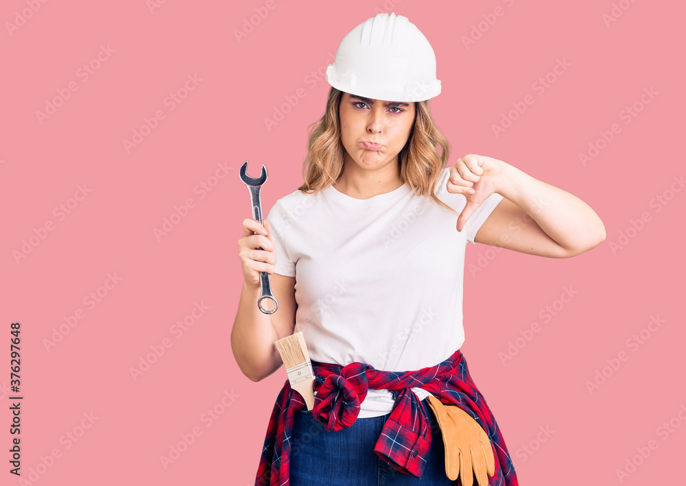 Young caucasian woman wearing hardhat holding wrench with angry face, negative sign showing dislike with thumbs down, rejection concept Young caucasian woman wearing hardhat holding wrench with angry face, negative sign showing dislike with thumbs down, rejection concept