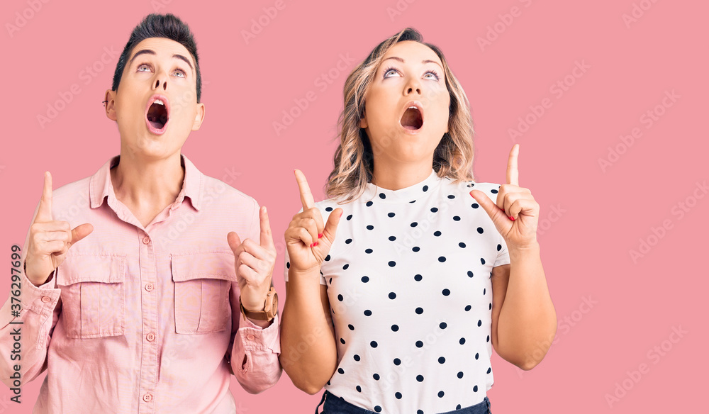 Couple of women wearing casual clothes amazed and surprised looking up and pointing with fingers and raised arms. Couple of women wearing casual clothes amazed and surprised looking up and pointing with fingers and raised arms.