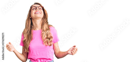 Young beautiful caucasian woman with blond hair wearing casual clothes and glasses very happy and excited doing winner gesture with arms raised, smiling and screaming for success. celebration concept.