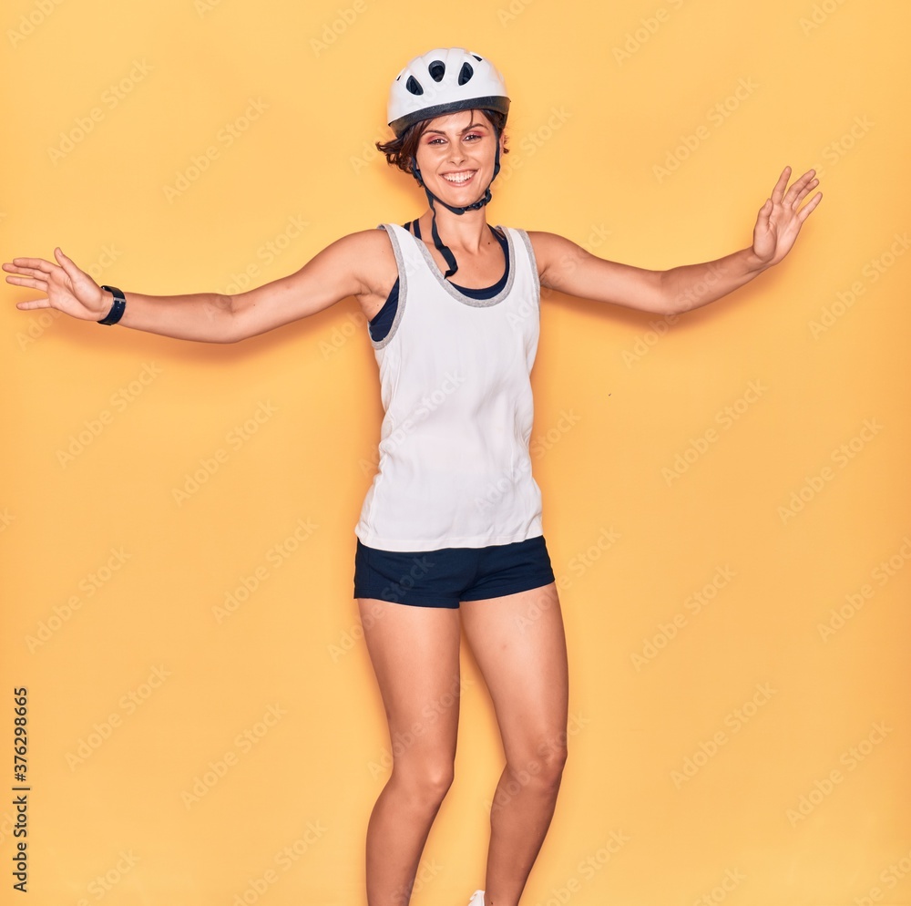 Young beautiful biker woman wearing bike helmet smiling happy. Jumping with smile on face and arms opened over isolated yellow background. Young beautiful biker woman wearing bike helmet smiling happy. Jumping with smile on face and arms opened over isolated yellow background.
