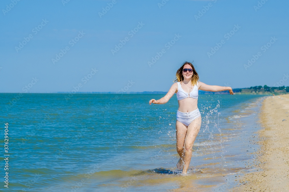 A young girl runs along the sandy sea beach along the surf. Splashes of sea water. The girl is wearing a white swimsuit and sunglasses. Freedom and carelessness. Outdoor fitness. Sunny day. Copy space A young girl runs along the sandy sea beach along the surf. Splashes of sea water. The girl is wearing a white swimsuit and sunglasses. Freedom and carelessness. Outdoor fitness. Sunny day. Copy space