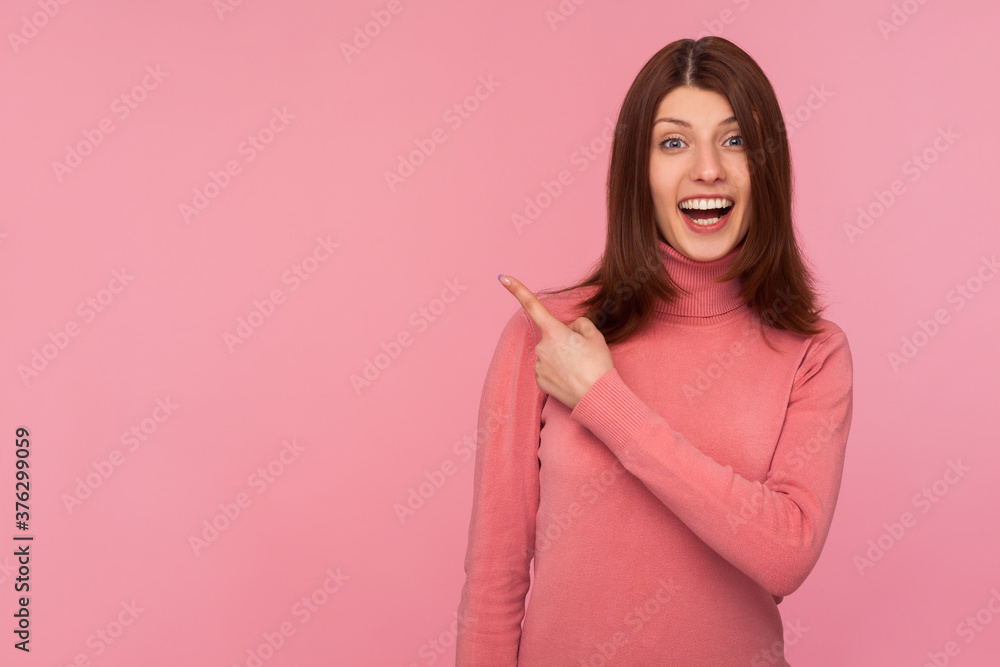 Obraz premium Happy surprised woman in pink sweater pointing finger away with toothy smile, wondering about empty space for your advertisement, template. Indoor studio shot isolated on pink background