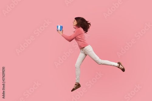 Full length portrait of smiling brunette girl jumping in air or flying with gift box, hurry to make holiday present. Indoor studio shot isolated on pink background