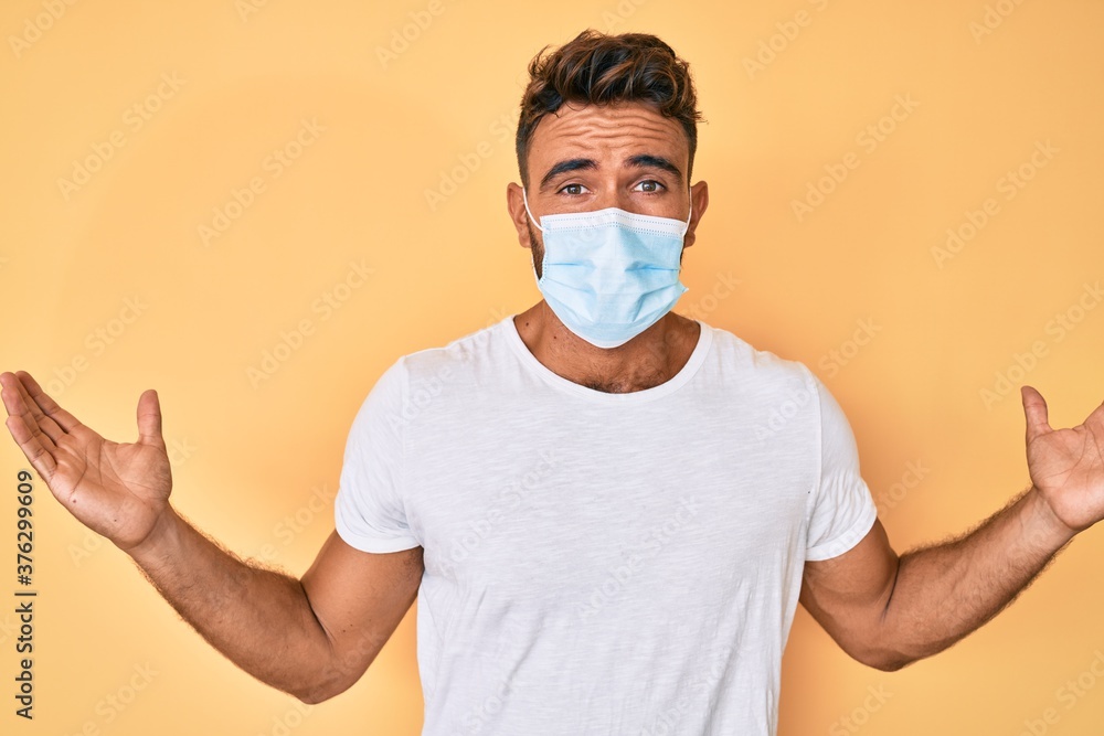 Young hispanic man wearing medical mask clueless and confused with open arms, no idea and doubtful face.