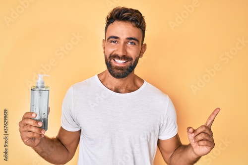 Young hispanic man using hand sanitizer gel smiling happy pointing with hand and finger to the side