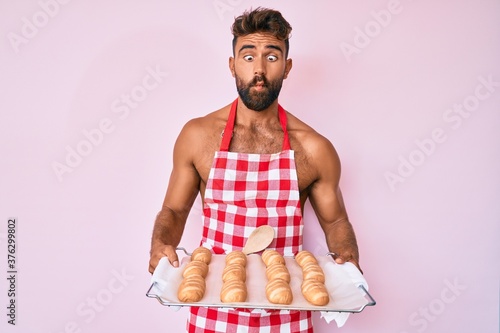 Young hispanic man shirtless wearing baker uniform holding homemade bread making fish face with mouth and squinting eyes, crazy and comical.