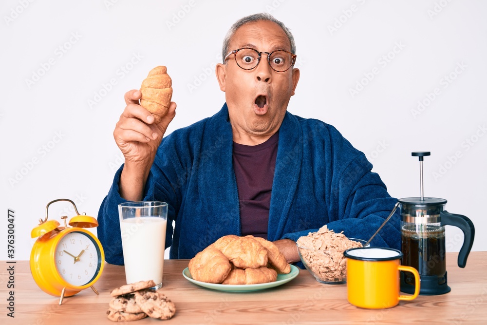 Senior handsome man with gray hair sitting on the table eating croissant for breakfast scared and amazed with open mouth for surprise, disbelief face Senior handsome man with gray hair sitting on the table eating croissant for breakfast scared and amazed with open mouth for surprise, disbelief face