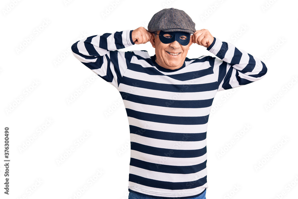 Senior handsome man wearing burglar mask and t-shirt smiling pulling ears with fingers, funny gesture. audition problem Senior handsome man wearing burglar mask and t-shirt smiling pulling ears with fingers, funny gesture. audition problem