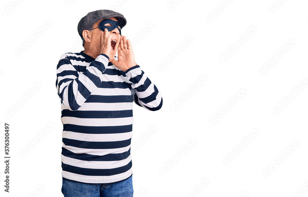 Senior handsome man wearing burglar mask and t-shirt shouting angry out loud with hands over mouth Senior handsome man wearing burglar mask and t-shirt shouting angry out loud with hands over mouth