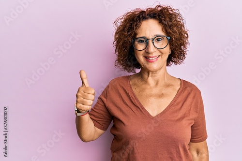 Beautiful middle age mature woman wearing casual clothes and glasses smiling happy and positive, thumb up doing excellent and approval sign