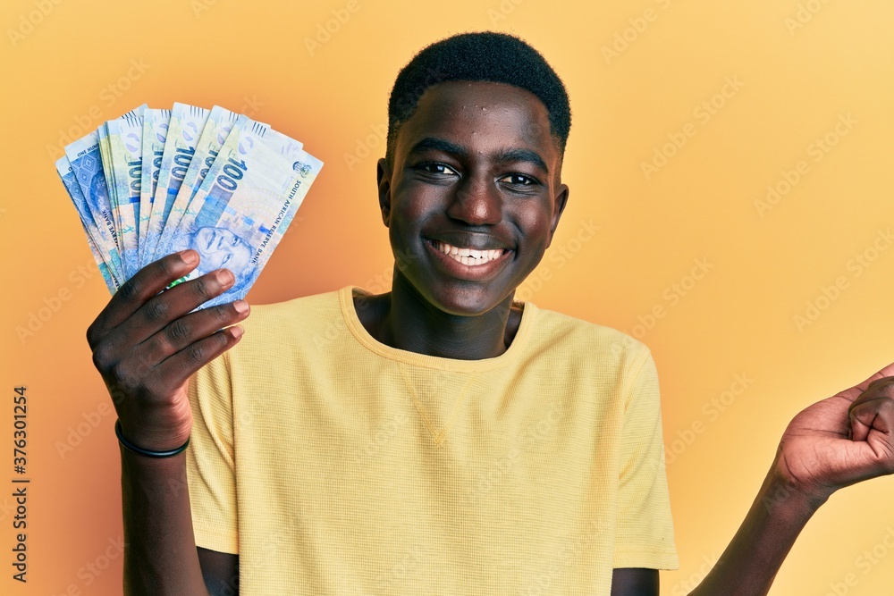 Young african american man holding south african rand banknotes screaming proud, celebrating victory and success very excited with raised arm Young african american man holding south african rand banknotes screaming proud, celebrating victory and success very excited with raised arm