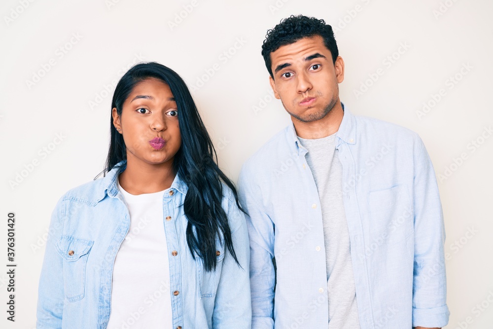 Beautiful latin young couple wearing casual clothes puffing cheeks with funny face. mouth inflated with air, crazy expression. Beautiful latin young couple wearing casual clothes puffing cheeks with funny face. mouth inflated with air, crazy expression.