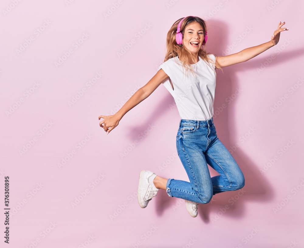 Young beautiful blonde woman smiling happy. Jumping with smile on face listening to music using pink headphones over isolated background Young beautiful blonde woman smiling happy. Jumping with smile on face listening to music using pink headphones over isolated background