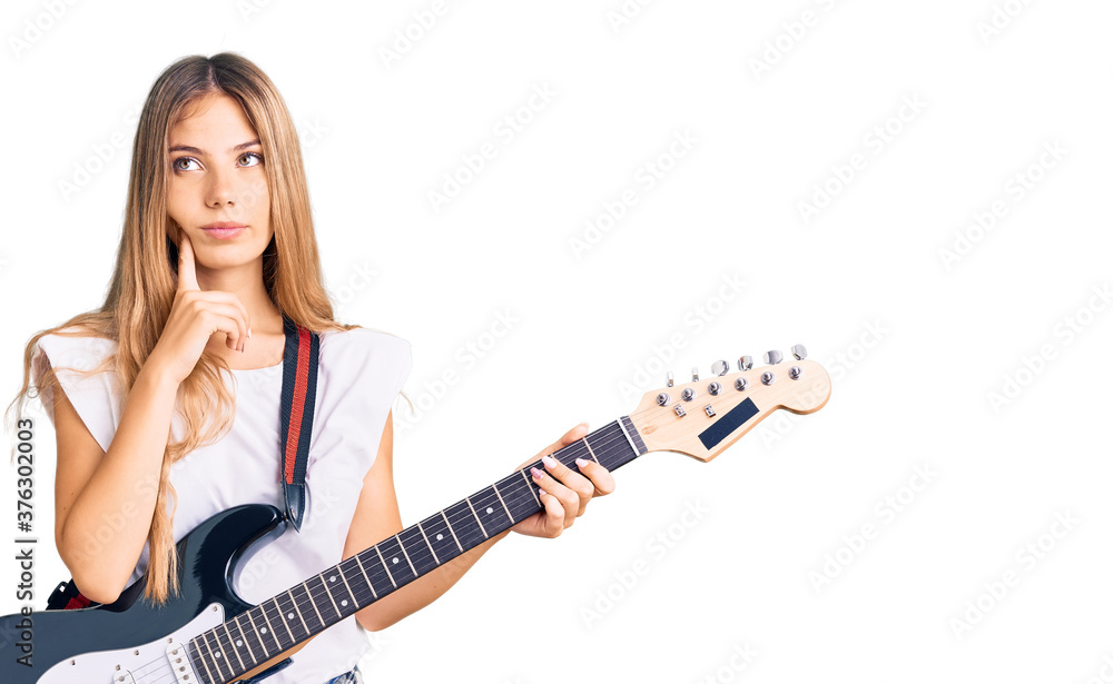 Beautiful caucasian woman with blonde hair playing electric guitar serious face thinking about question with hand on chin, thoughtful about confusing idea