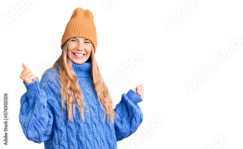 Beautiful caucasian woman with blonde hair wearing wool sweater and winter hat very happy and excited doing winner gesture with arms raised, smiling and screaming for success. celebration concept. photo