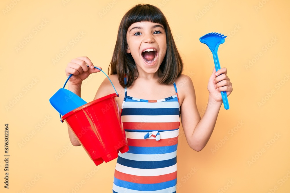 Young little girl with bang playing with summer shovel and bucket toys celebrating crazy and amazed for success with open eyes screaming excited. Young little girl with bang playing with summer shovel and bucket toys celebrating crazy and amazed for success with open eyes screaming excited.