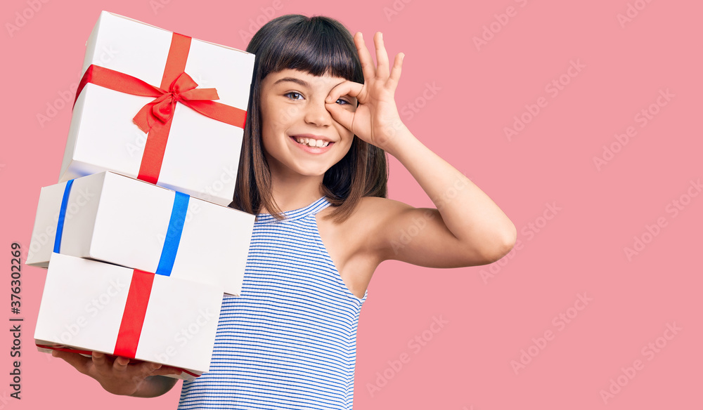 Young little girl with bang holding gifts smiling happy doing ok sign with hand on eye looking through fingers Young little girl with bang holding gifts smiling happy doing ok sign with hand on eye looking through fingers