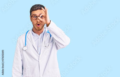 Handsome young man with bear wearing doctor uniform doing ok gesture shocked with surprised face, eye looking through fingers. unbelieving expression.