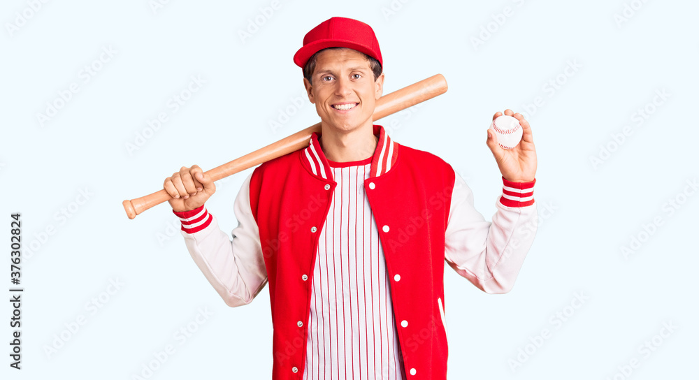 Young handsome man playing baseball holding bat and ball looking positive and happy standing and smiling with a confident smile showing teeth Young handsome man playing baseball holding bat and ball looking positive and happy standing and smiling with a confident smile showing teeth