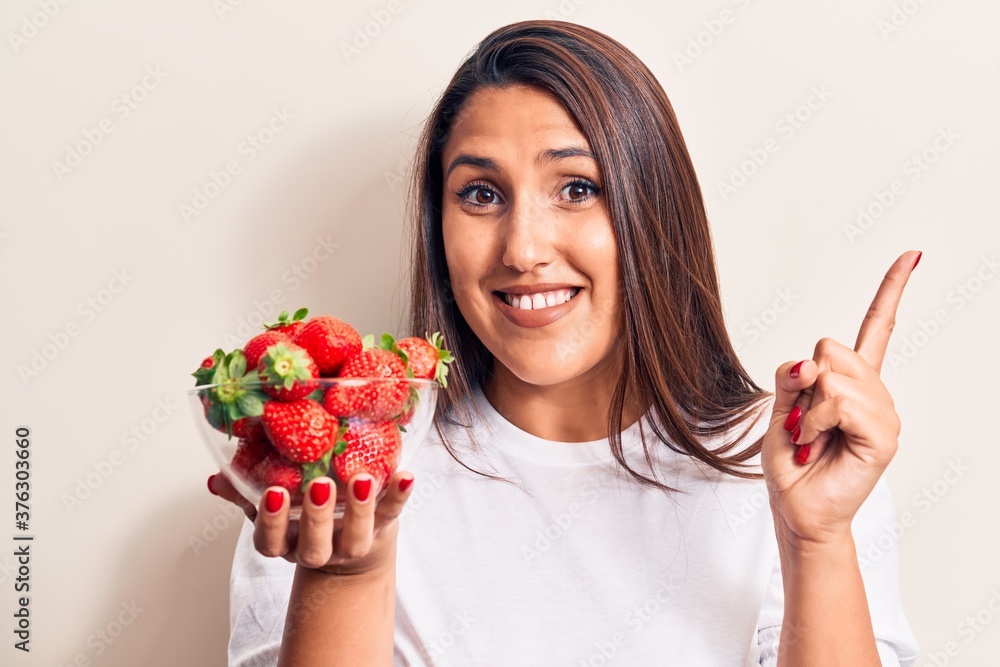 Young beautiful brunette woman holding bowl with strawberries smiling happy pointing with hand and finger to the side