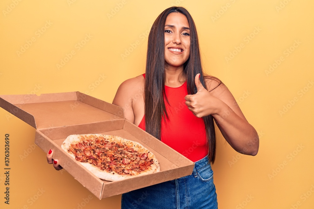 Young beautiful brunette woman holding delivery box with italian pizza smiling happy and positive, thumb up doing excellent and approval sign