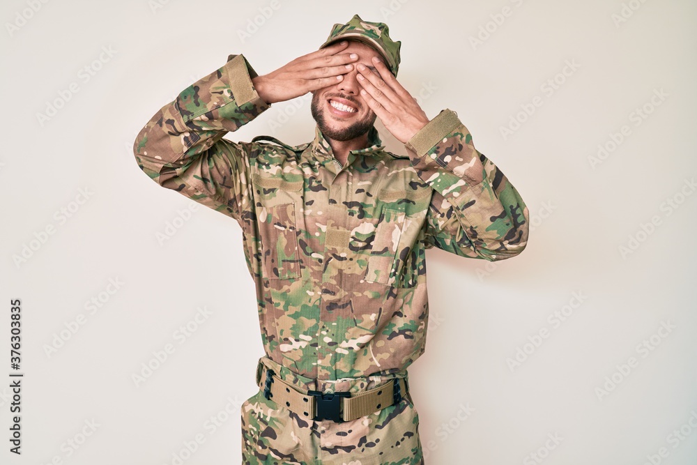 Fototapeta premium Young caucasian man wearing camouflage army uniform covering eyes with hands smiling cheerful and funny. blind concept.