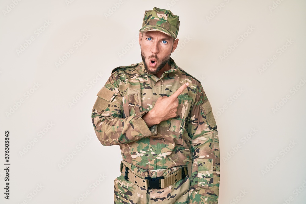 Young caucasian man wearing camouflage army uniform surprised pointing with finger to the side, open mouth amazed expression. Young caucasian man wearing camouflage army uniform surprised pointing with finger to the side, open mouth amazed expression.