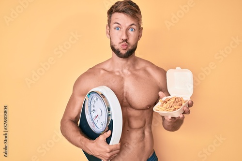 Young caucasian man shirtless holding weighing machine and fried potatoes puffing cheeks with funny face. mouth inflated with air  catching air.