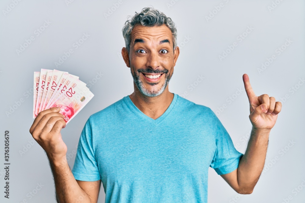Middle age grey-haired man holding israel shekels smiling with an idea or question pointing finger with happy face, number one