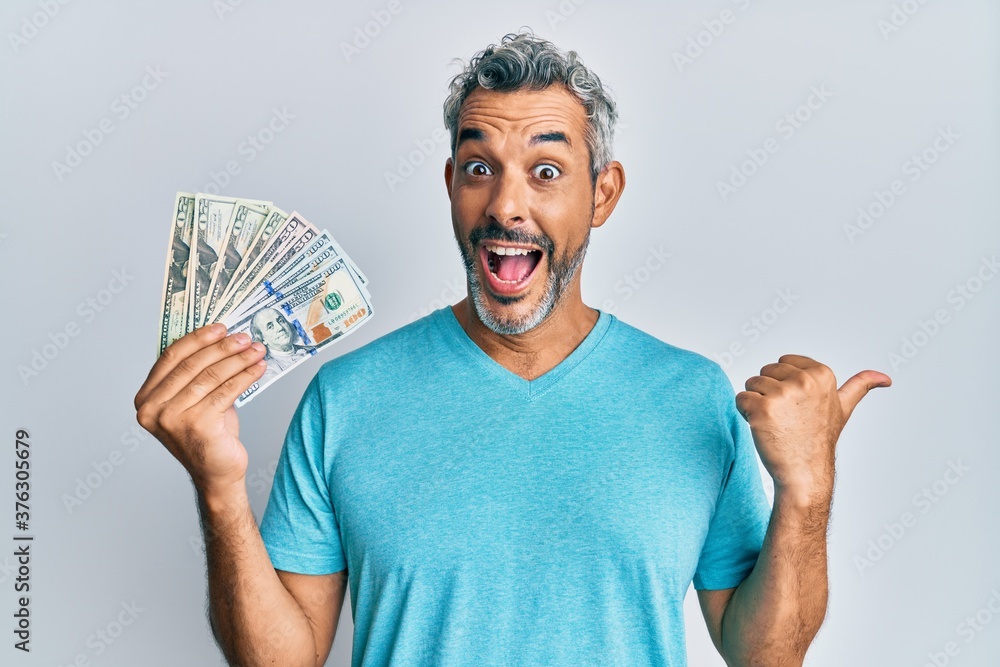 Middle age grey-haired man holding dollars pointing thumb up to the side smiling happy with open mouth Middle age grey-haired man holding dollars pointing thumb up to the side smiling happy with open mouth