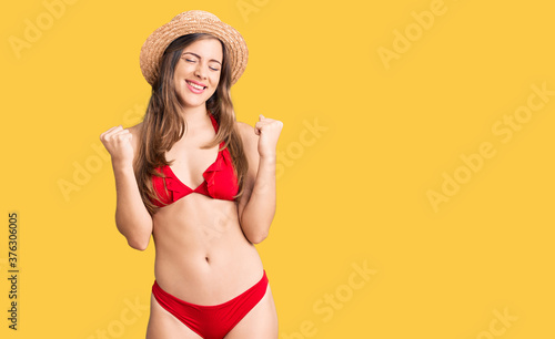 Beautiful caucasian young woman wearing bikini and summer hat very happy and excited doing winner gesture with arms raised, smiling and screaming for success. celebration concept.