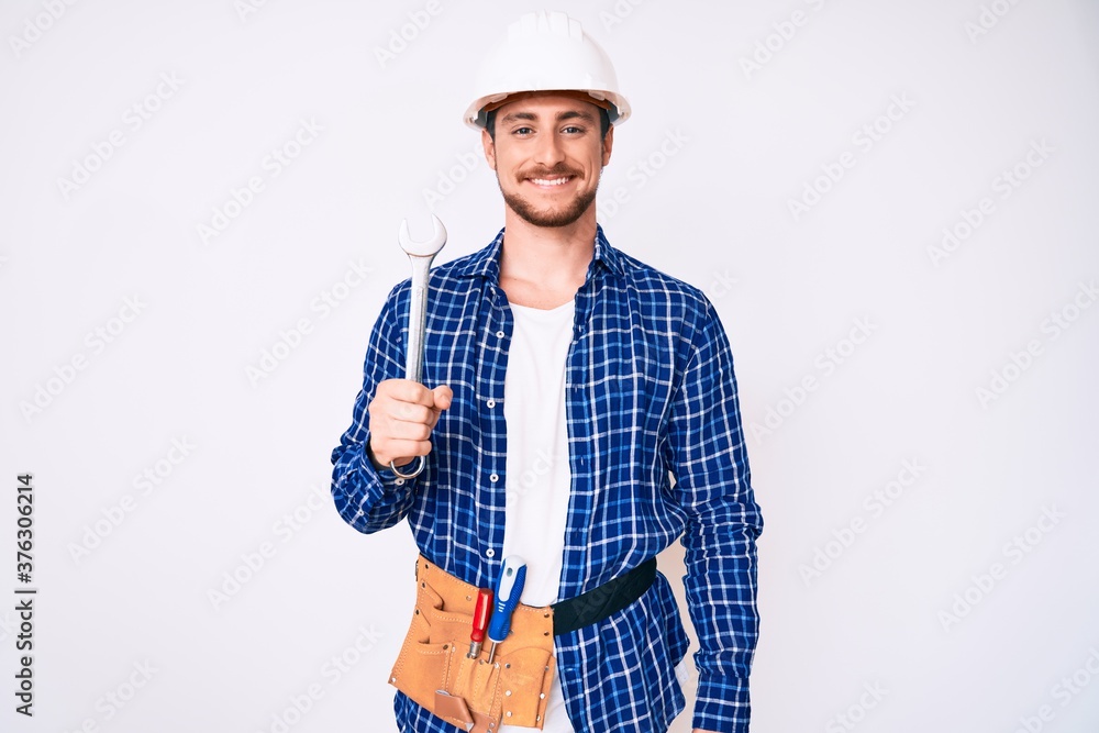 Young handsome man weaing handyman uniform holding wrench looking positive and happy standing and smiling with a confident smile showing teeth Young handsome man weaing handyman uniform holding wrench looking positive and happy standing and smiling with a confident smile showing teeth