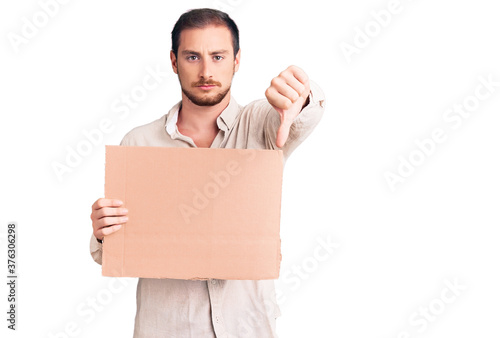 Young handsome caucasian man holding empty cardboard banner with angry face, negative sign showing dislike with thumbs down, rejection concept