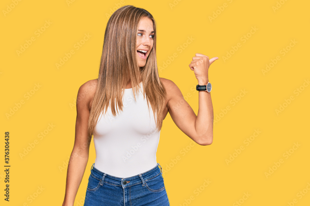 Young blonde woman wearing casual style with sleeveless shirt smiling with happy face looking and pointing to the side with thumb up. Young blonde woman wearing casual style with sleeveless shirt smiling with happy face looking and pointing to the side with thumb up.