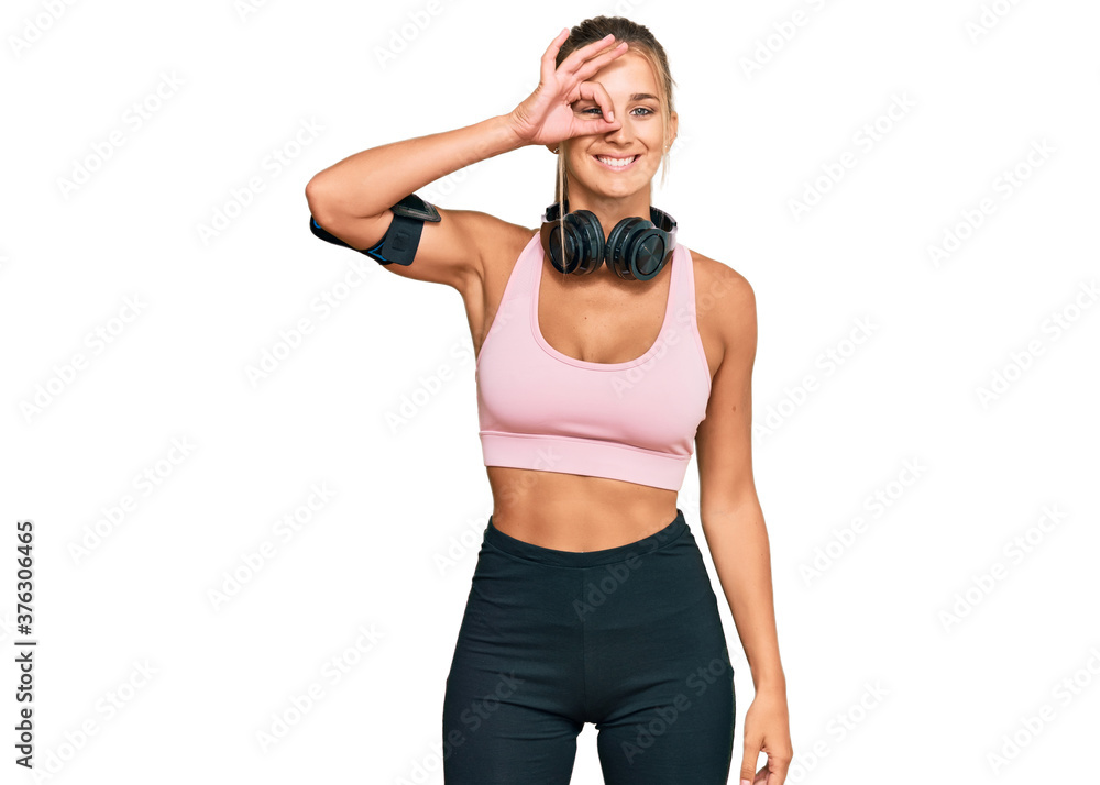 Young blonde woman wearing gym clothes and using headphones doing ok gesture with hand smiling, eye looking through fingers with happy face. Young blonde woman wearing gym clothes and using headphones doing ok gesture with hand smiling, eye looking through fingers with happy face.