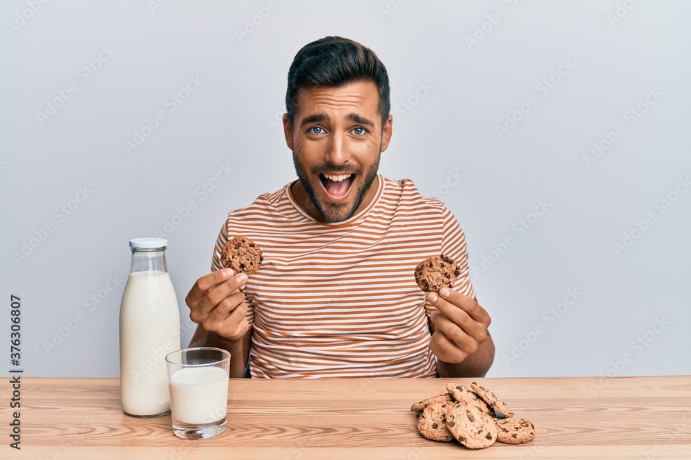 Handsome hispanic man drinking a glass of milk holding chocolate cookies smiling and laughing hard out loud because funny crazy joke. Handsome hispanic man drinking a glass of milk holding chocolate cookies smiling and laughing hard out loud because funny crazy joke.