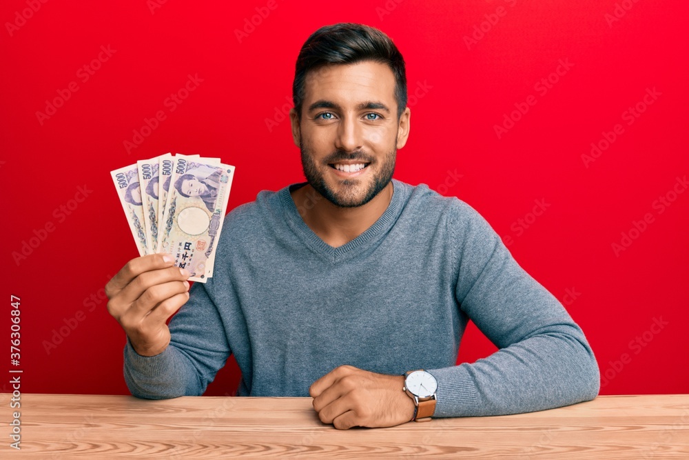 Handsome hispanic man holding japanese yen banknotes looking positive and happy standing and smiling with a confident smile showing teeth Handsome hispanic man holding japanese yen banknotes looking positive and happy standing and smiling with a confident smile showing teeth